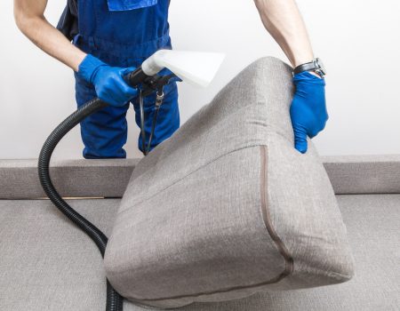 Cleaning service. Man janitor in gloves and uniform vacuum clean sofa with professional equipment.